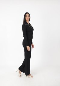 Schwarze gerippte Zip-Jacke mit Kragen über schwarzen schmalen Hosen. Getragen mit weißen Absatzsandalen. Einfaches Design und taillierte Silhouette.