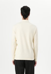 Polo à manches longues de couleur crème, fabriqué en coton doux, avec un col classique et des poignets côtelés. Design uni, détails minimaux.