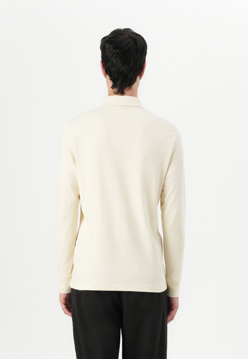 Langarm-Polohemd in Cremefarbe, aus weicher Baumwolle gefertigt, mit klassischem Kragen und gerippten Bündchen. Schlichtes Design, minimalistische Details.