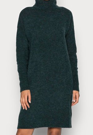 Ciemnozielona, dzianinowa sukienka z golfem i długim rękawem, noszona przez osobę z widocznymi dłońmi i nogami na jednolitym tle.