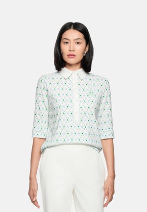 Frau trägt ein weißes Hemd mit Kragen, verziert mit einem Muster aus grünen und blauen Herzen und halblangen Ärmeln, kombiniert mit weißen High-Waist-Hosen, steht vor einem einfarbigen Hintergrund.