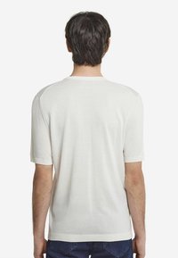 Vit stickad kortärmad tröja med rund hals, som har en slät textur och ribbade ärmslut, sedd från baksidan.