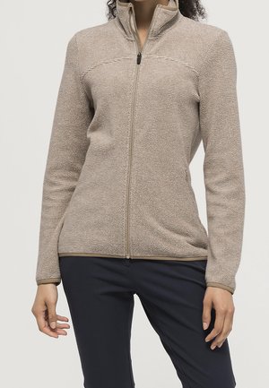 Kvinde iført beige tekstureret lynlåstrøje og mørke bukser, stående med hænder afslappet ned langs siden.