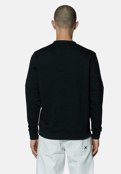 Felpa nera a girocollo con maniche lunghe, polsini e orlo a coste; presenta una texture di cotone liscia. Indossata con pantaloni bianchi.