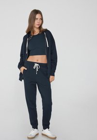 Sudadera azul marino con cordón blanco, combinada con joggers a juego y un top corto. Los zapatos son blancos con una suela marrón clara.