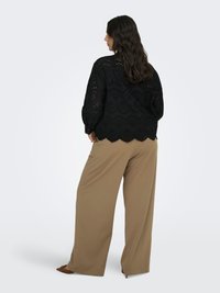 Vrouw met lang donker haar, gekleed in een zwarte eyeletblouse en beige wijde broek, staand en iets weggedraaid tegen een effen achtergrond.