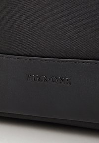 Svart veske med strukturert stoffoverside og glatt lærbunn, preget logodetalj, rektangulær form og minimalistisk design.
