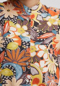 Chemise à boutons multicolore à motifs floraux, présentant de grandes fleurs éclatantes en jaune, orange et bleu sur un fond sombre ; tissu texturé.