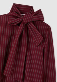 Camicia color borgogna con righe bianche verticali, caratterizzata da un grande fiocco al collo e tessuto liscio e strutturato.