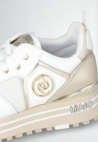 Zapatilla blanca y dorada con parte superior texturizada, detalle del logo en dorado y suela gruesa beige con rayas blancas. Los acentos incluyen elementos metálicos.