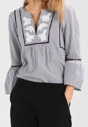 Mujer con blusa de rayas verticales en blanco y negro con bordado floral blanco y pantalones negros, manos en los bolsillos.