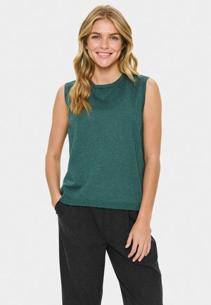 Smiling woman with wavy blonde hair wearing a sleeveless green top and loose black pants, standing with one hand in pocket.
