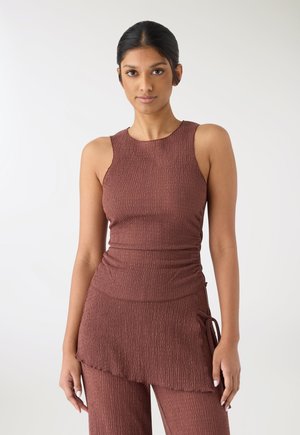 Donna in piedi su uno sfondo bianco, indossa un top senza maniche strutturato e pantaloni coordinati marrone tenue con dettaglio annodato sul lato.