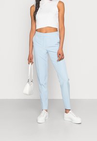 Ljust blå skräddarsydda byxor med en slät textur, kombinerat med en vit ribbad crop top och vita platåskor. Håller i en liten vit väska.