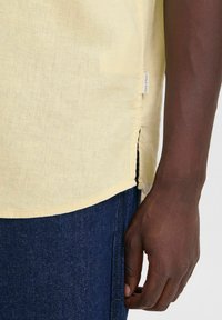 Camicia di lino gialla con spacchi laterali, orlo curvo e un'etichetta sulla cucitura, indossata sopra pantaloni di denim blu.