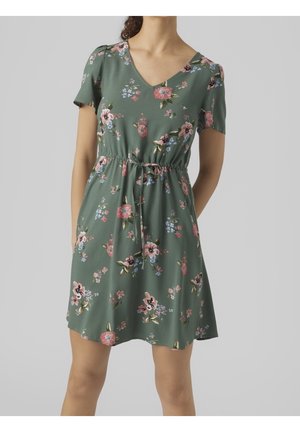 Femme portant une robe courte verte à fleurs avec col en V et ceinture à la taille, debout devant un fond uni gris clair.