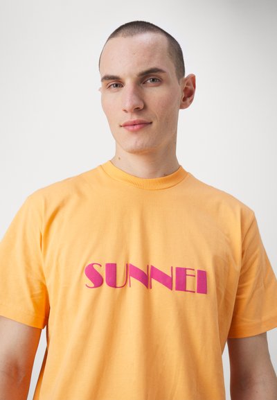 Oransje bomulls T-skjorte med korte ermer, med en iøynefallende rosa trykk som staver "SUNNEI" på forsiden. Enkel design, myk tekstur.