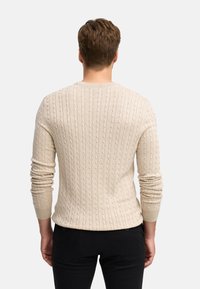 Maglione a maglia beige con un motivo a treccia testurizzato, maniche lunghe e scollatura rotonda, abbinato a pantaloni scuri aderenti.