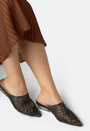 Füße mit schwarzen, spitz zulaufenden Slip-On-Schuhen aus gewebtem Mesh, kombiniert mit einem braunen, plissierten Rock auf einem einfarbigen hellgrauen Hintergrund.