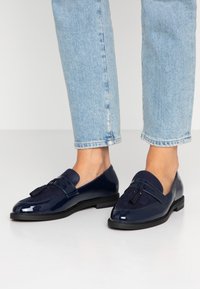 Mørkeblå loafers i blankt lakkmateriale, med suedepreg, og en duskdetalj, kombinert med lyseblå croppede jeans.