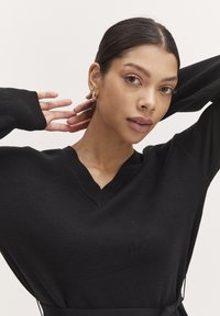 Maglione nero a maglia con scollatura a V, maniche lunghe e vita aderente. Texture liscia e design minimalista, indossato da un modello.