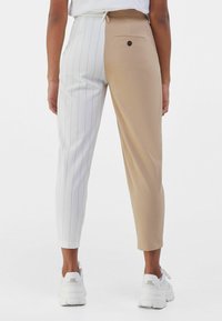 Byxor med en delad design: det ena benet är beige, det andra vitt med smala ränder. Har en bakficka och knappstängning. Vita sneakers.