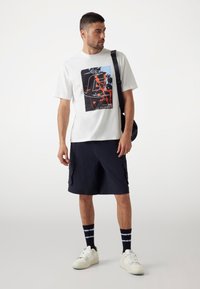 Camiseta de algodón blanca con un estampado gráfico en negro y rojo, combinada con pantalones cortos cargo azul marino, calcetines a rayas en blanco y negro, y zapatillas blancas.