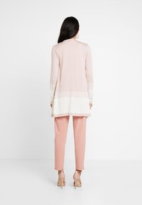 Ljusrosa lång kofta med vita horisontella ränder i nederkant och ärmslut, i kombination med smala rosa byxor och beige höga klackar.