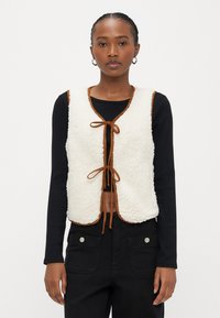 Hvit fleecevest med brun kant, med v-hals og knyting foran. Brukt over en svart langermet topp og svarte bukser.