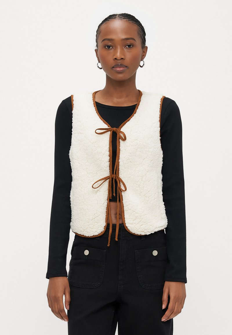 Hvit fleecevest med brun kant, med v-hals og knyting foran. Brukt over en svart langermet topp og svarte bukser.