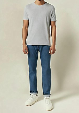 Mand iført en lysegrå kortærmet t-shirt, blå opsmøgede jeans og hvide sneakers, stående mod en ensfarvet baggrund.