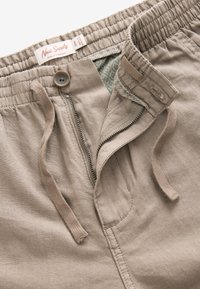 Pantaloni corti beige con vita elastica dotata di cordoncino, zip anteriore e fodera interna a fantasia. Etichetta con la scritta "Next Supply."
