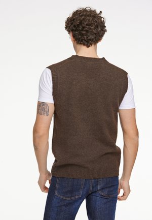 Bruine gebreide mouwloze trui met een geribde zoom, gedragen over een wit shirt, gecombineerd met blauwe jeans. Achteraanzicht toont textuur en pasvorm.
