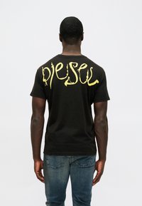 Vyras vilkintis juodą marškinėlį su geltonai stilizuotu tekstu "diesel" ant nugaros ir mėlynas džinsus, stovi prieš baltą sieną.