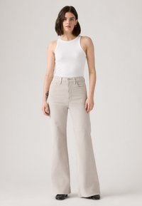 Calças de ganga bege claro com corte flare e cintura alta. Apresentam bolsos à frente, passantes para cinto e fecho com botão. Combinadas com uma t-shirt branca sem mangas.
