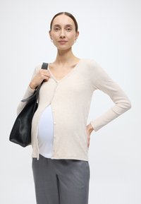 Μπεζ ριγωτή εγκυμοσυνητική ζακέτα με μακριά μανίκια και κουμπιά μπροστά, φορεμένη πάνω από λευκό τοπ. Το μοντέλο κρατάει μαύρη δερμάτινη τσάντα, συνδυασμένη με γκρι παντελόνι.