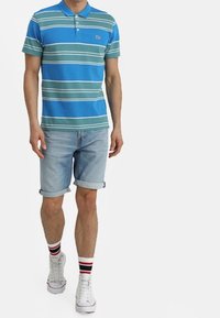 Blå och grönrandig polo tröja med krage, i kombination med uppvikta ljusblå denimshorts och vita sneakers med röda och svarta ränder på strumporna.