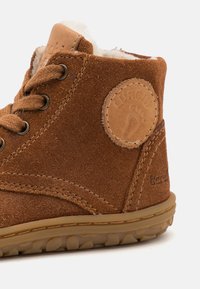 Hnědé semišové kotníkové boty s texturovaným povrchem, přední část s kulatou koženou nášivkou a pevnými šněrováním. Flexibilní gumová podrážka s vzorovaným profilem.