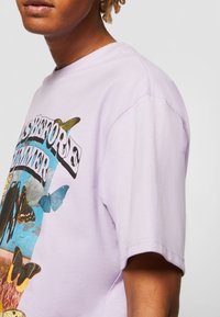 Tricou din bumbac lavandă, cu guler rotund și mâneci scurte, având grafice vibrante cu fluturi și textul "ESTE ÎNAINTE DE VARĂ."