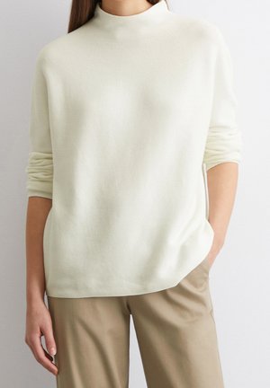 Femme portant un pull crème à manches longues avec les poignets retroussés et un pantalon beige, debout devant un fond clair uni.