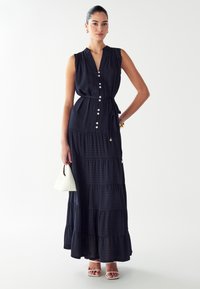 Námornícky maxi šaty so stupňovým strihom, V-výstrihom a bez rukávov, s gombíkovým zapínaním vpredu a tkanicou na obvazovanie v páse. Hladká textúra, doplnené o bielu kabelku.