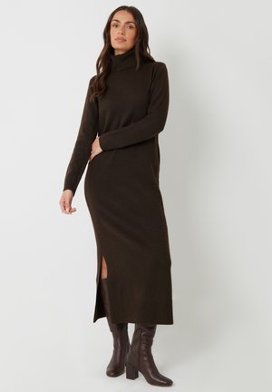 Langes braunes Strickkleid mit hohem Ausschnitt und Seitenschlitzen, kombiniert mit dunkelbraunen Absatzstiefeln, die ein tailliertes Design und gerippte Textur aufweisen.