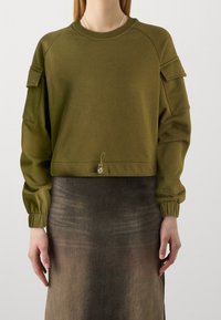 Personne portant un sweat-shirt court à manches longues vert olive avec des poches sur les manches et une jupe ombrée foncée.
