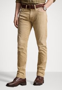 Pantalones de algodón beige con corte ajustado, diseño de cinco bolsillos, combinados con botas de cuero marrón y un cinturón texturizado a juego.