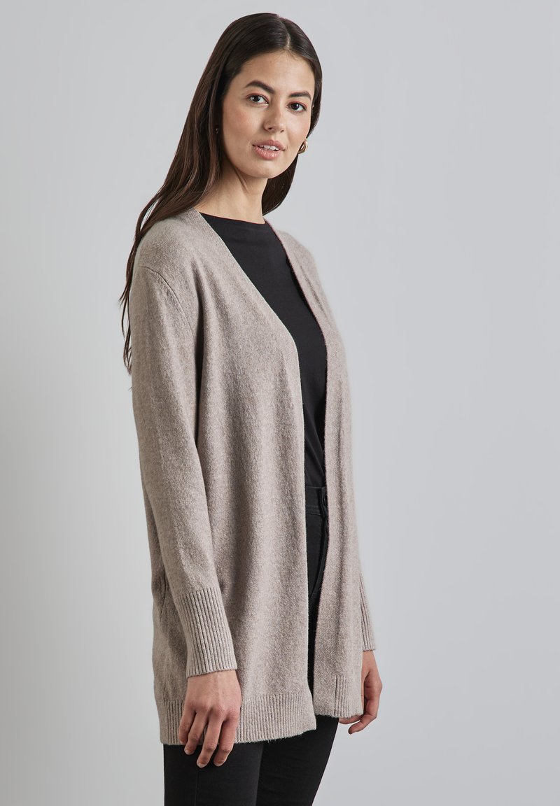 Cardigan ouvert beige clair en mélange de laine douce, présentant une coupe décontractée, des poignets côtelés et une texture lisse. Porté sur un haut noir.