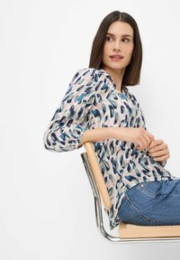 Durchsichtige Bluse in Hellrosa mit blauen und grünen abstrakten Mustern, langen Ärmeln, Kragen und einer lockeren Passform, kombiniert mit Jeans.