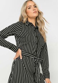 Schwarz-weiß gestreifte Bluse mit langen Ärmeln, Kragen und gebundenem Taillendetail. Merkmale hervorstehende vertikale Streifen und zwei Taschen.