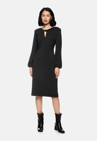 Schwarzes Kleid mit langen Puffärmeln, Schlitzdetail am Ausschnitt, taillierter Silhouette und knieumspielender Länge. Getragen mit schwarzen Absatzstiefeln.