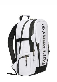 Sac à dos blanc avec des accents noirs, présentant de multiples poches zippées, des sangles réglables et un logo "SUPERDRY" bien en vue. Tissu durable.