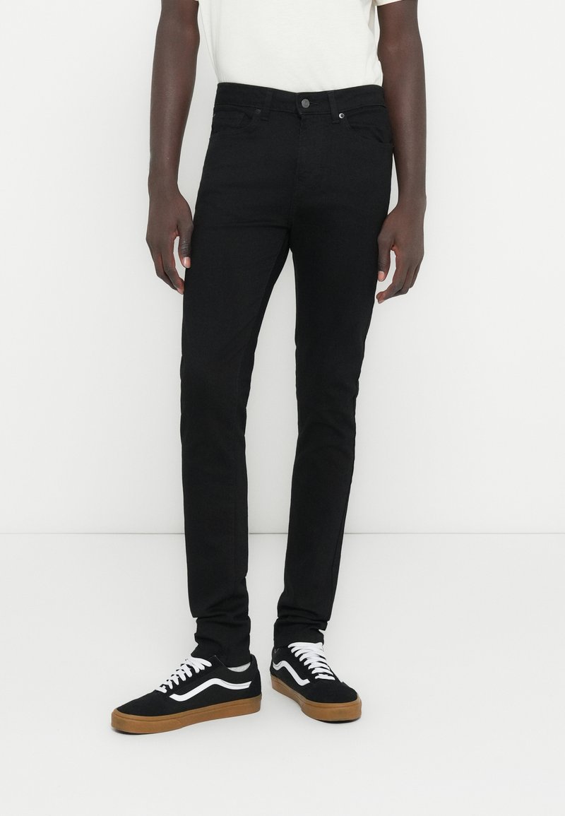 Vaqueros pitillo negros hechos de denim, con un diseño clásico de cinco bolsillos, textura suave y cierre de botón. Combinados con zapatillas negras y blancas.